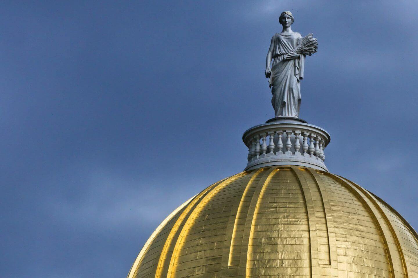 Gold leafed dome of the Vermont State House against blue sky with carved wooden statue of the Goddess Ceres.